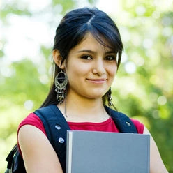 A girl with dark hair smiles at the camera while wearing a backpack and holding a book. 